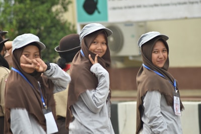 Three young women wearing uniforms with brown hijabs and gray caps are smiling and posing for the camera. Two of them make peace signs, and they have lanyards with ID cards around their necks. The setting appears to be an outdoor event, possibly linked to scouts or a similar organization. Behind them is a blurred background with greenery and a building with an air conditioning unit.