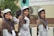 Three young women wearing uniforms with brown hijabs and gray caps are smiling and posing for the camera. Two of them make peace signs, and they have lanyards with ID cards around their necks. The setting appears to be an outdoor event, possibly linked to scouts or a similar organization. Behind them is a blurred background with greenery and a building with an air conditioning unit.