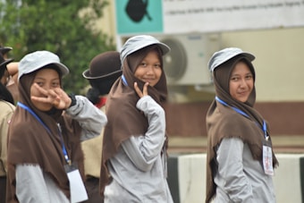 Three young women wearing uniforms with brown hijabs and gray caps are smiling and posing for the camera. Two of them make peace signs, and they have lanyards with ID cards around their necks. The setting appears to be an outdoor event, possibly linked to scouts or a similar organization. Behind them is a blurred background with greenery and a building with an air conditioning unit.
