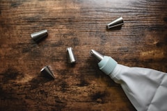 On a wooden surface, several metal icing tips are scattered alongside a white piping bag with a teal coupler.