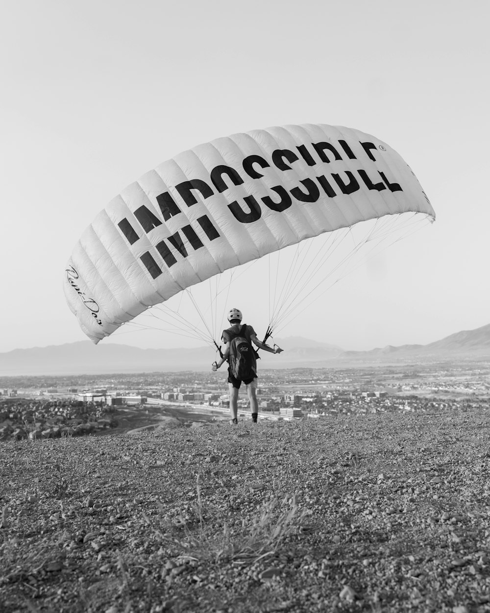 Foto en escala de grises de un hombre con camisa negra y pantalones sosteniendo un paracaídas