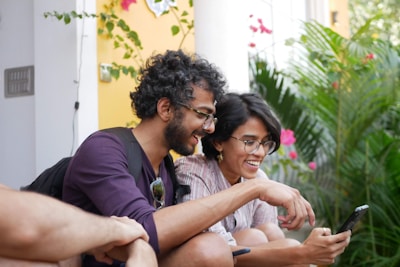Two people sitting outside, smiling while looking at a smartphone. They appear to be enjoying a moment together surrounded by greenery and a vibrant yellow wall. Their expressions suggest they are engaged in a pleasant conversation or viewing something amusing.