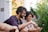 Two people sitting outside, smiling while looking at a smartphone. They appear to be enjoying a moment together surrounded by greenery and a vibrant yellow wall. Their expressions suggest they are engaged in a pleasant conversation or viewing something amusing.