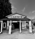 A rustic general store with a wooden porch and signs advertising coffee, pickles, and other goods. Two wooden chairs are placed on either side of the entrance. The facade features a vintage design with a prominent sign labeled 'Rust General Store'.