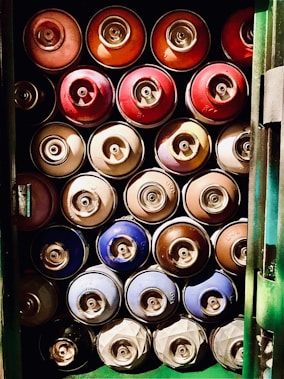 A collection of spray paint cans arranged in a neat grid pattern inside a storage container. The cans are of various colors, including red, orange, blue, and brown. They are stacked in four rows and three columns, with the caps facing outward.