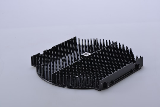 A black, circular object with an intricate pattern of evenly spaced vertical pins and a square metallic component at the center. The object appears to have a segmented design with sections of different heights.