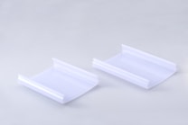 Two identical clear plastic items are positioned side by side on a smooth, light gray surface. Each item has a curved, rectangular shape with raised edges on two opposite sides.