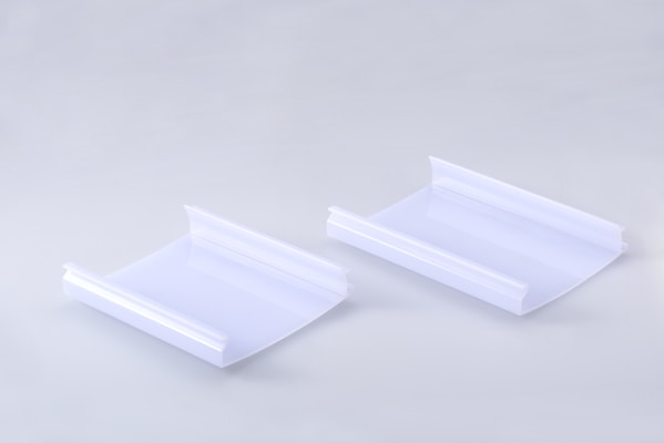 Two identical clear plastic items are positioned side by side on a smooth, light gray surface. Each item has a curved, rectangular shape with raised edges on two opposite sides.
