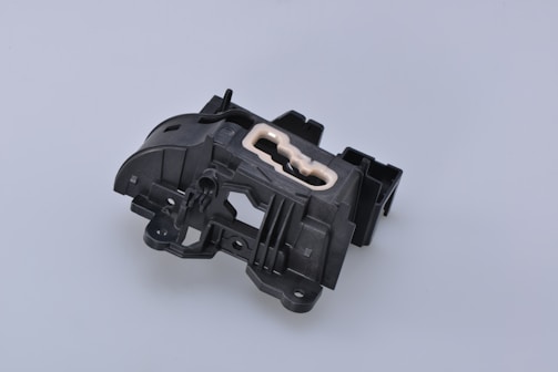 A complex black plastic mechanical component with a beige insert in the center. The piece features various geometric shapes and cutouts, suggesting it might be part of a larger machinery or device.