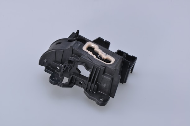 A complex black plastic mechanical component with a beige insert in the center. The piece features various geometric shapes and cutouts, suggesting it might be part of a larger machinery or device.
