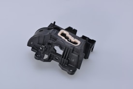 A complex black plastic mechanical component with a beige insert in the center. The piece features various geometric shapes and cutouts, suggesting it might be part of a larger machinery or device.