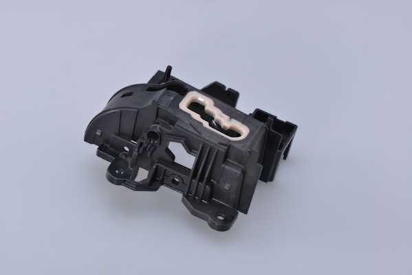 A complex black plastic mechanical component with a beige insert in the center. The piece features various geometric shapes and cutouts, suggesting it might be part of a larger machinery or device.