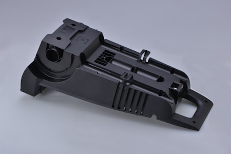 A complex, black plastic component with various ridges and openings, likely part of a larger mechanical structure. The item has a rectangular base with several precise cutouts and a cylindrical opening, suggesting a functional purpose.