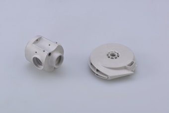 Two white mechanical components are positioned on a plain light gray background. The component on the left has a cylindrical shape with multiple threaded holes and a few visible screws, suggesting a part of a larger assembly or system. The component on the right is circular and flat with a central hollow area, possibly a spool or housing for internal mechanisms.