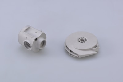 Two white mechanical components are positioned on a plain light gray background. The component on the left has a cylindrical shape with multiple threaded holes and a few visible screws, suggesting a part of a larger assembly or system. The component on the right is circular and flat with a central hollow area, possibly a spool or housing for internal mechanisms.
