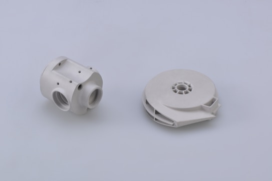 Two white mechanical components are positioned on a plain light gray background. The component on the left has a cylindrical shape with multiple threaded holes and a few visible screws, suggesting a part of a larger assembly or system. The component on the right is circular and flat with a central hollow area, possibly a spool or housing for internal mechanisms.