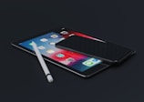 A sleek tablet lies on a dark surface, displaying a colorful home screen with various app icons. A smartphone rests on top of the tablet, its screen turned off. Beside them is a white stylus, positioned diagonally across the tablet.