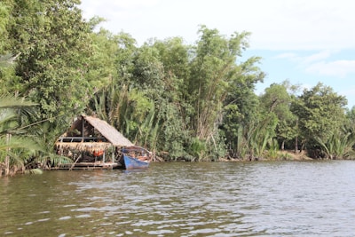 A wooden hut with a thatched roof nestles among dense green foliage along the banks of a calm river. A small blue boat is moored adjacent to the hut, partially sheltered by hanging palm leaves. The scene is serene and enveloped by abundant greenery.