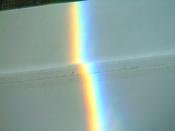 A bright, vivid rainbow reflection appears on a light-colored surface. The colors transition smoothly from red on the left to violet on the right, creating a striking contrast against the plain background.