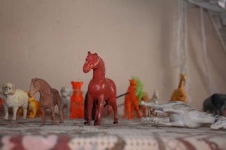 A collection of small plastic animal figurines is displayed on a surface. The figures include various animals such as horses, a sheep, a lion, and possibly other creatures. They are arranged in a row with some figures standing upright and one figure lying on its side. The colors of the figurines are varied, featuring red, brown, white, and yellow hues. The background is plain and neutral, with a focus on the figurines.