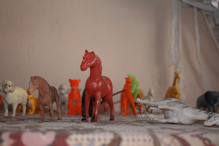 A collection of small plastic animal figurines is displayed on a surface. The figures include various animals such as horses, a sheep, a lion, and possibly other creatures. They are arranged in a row with some figures standing upright and one figure lying on its side. The colors of the figurines are varied, featuring red, brown, white, and yellow hues. The background is plain and neutral, with a focus on the figurines.