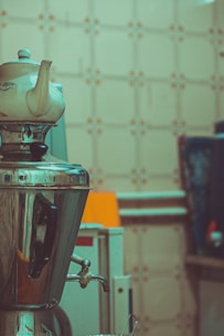 A ceramic teapot sits atop a shiny, metallic samovar, reflecting the tiled kitchen wall in the background. The scene suggests an old-fashioned kitchen or tea-making setup.