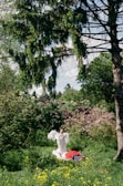 A person dressed in a white gown with large angel wings is standing in a lush green garden surrounded by blooming flowers and tall trees. A vibrant red blanket is spread out on the grass, with yellow wildflowers scattered around.
