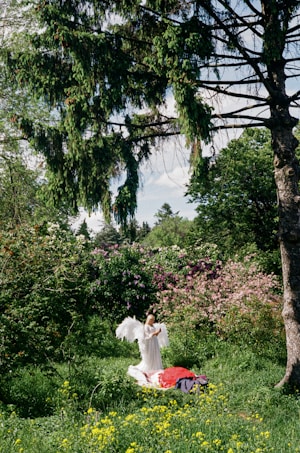 A person dressed in a white gown with large angel wings is standing in a lush green garden surrounded by blooming flowers and tall trees. A vibrant red blanket is spread out on the grass, with yellow wildflowers scattered around.