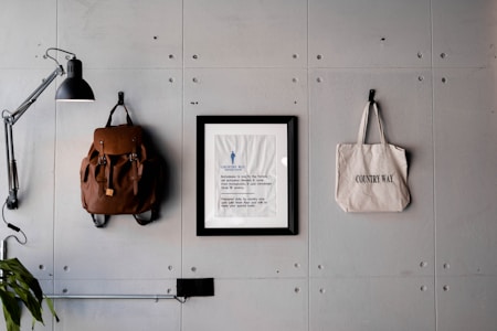 A minimalist wall arrangement features a modern black lamp, a brown leather backpack, a framed print with text, and a canvas tote bag labeled 'Country Way.' The objects are organized neatly against a textured gray wall with visible bolts, contributing to an industrial aesthetic.