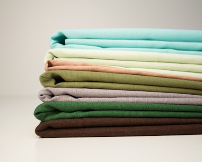A stack of neatly folded fabrics in various shades of green, brown, and beige is placed horizontally on a smooth surface. The materials appear to be made of linen or a similar textured fabric, arranged in a gradient from bright aqua and light mint to earthy, darker tones.