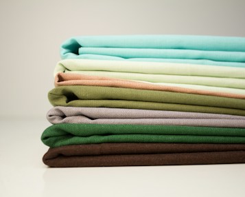 A stack of neatly folded fabrics in various shades of green, brown, and beige is placed horizontally on a smooth surface. The materials appear to be made of linen or a similar textured fabric, arranged in a gradient from bright aqua and light mint to earthy, darker tones.