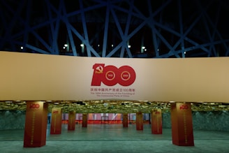A well-lit exhibition space features large pillars and banners celebrating the 100th Anniversary of the founding of the Communist Party of China. The design includes prominent red and gold elements, with the number 100 and the years 1921 and 2021. The background shows angular architectural structures with a modern aesthetic.