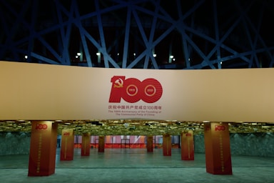 A well-lit exhibition space features large pillars and banners celebrating the 100th Anniversary of the founding of the Communist Party of China. The design includes prominent red and gold elements, with the number 100 and the years 1921 and 2021. The background shows angular architectural structures with a modern aesthetic.