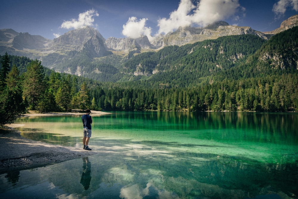 homem de camiseta preta e shorts azuis em pé na rocha perto do lago durante o dia