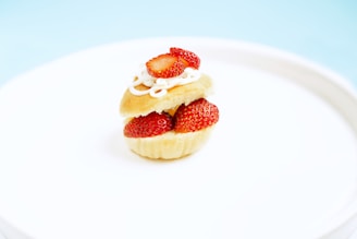 A small, layered dessert consisting of a sliced vanilla cupcake filled with fresh strawberry slices and topped with whipped cream and a strawberry slice. It is placed on a pristine white plate against a light blue background.