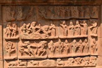 A detailed stone carving depicting a variety of figures engaged in different activities. The carving features multiple panels with figures in dynamic poses and expressions, capturing a sense of movement and storytelling. The figures appear to be dressed in traditional attire and some are adorned with headgear or ornate jewelry.
