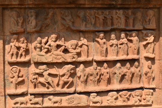 A detailed stone carving depicting a variety of figures engaged in different activities. The carving features multiple panels with figures in dynamic poses and expressions, capturing a sense of movement and storytelling. The figures appear to be dressed in traditional attire and some are adorned with headgear or ornate jewelry.