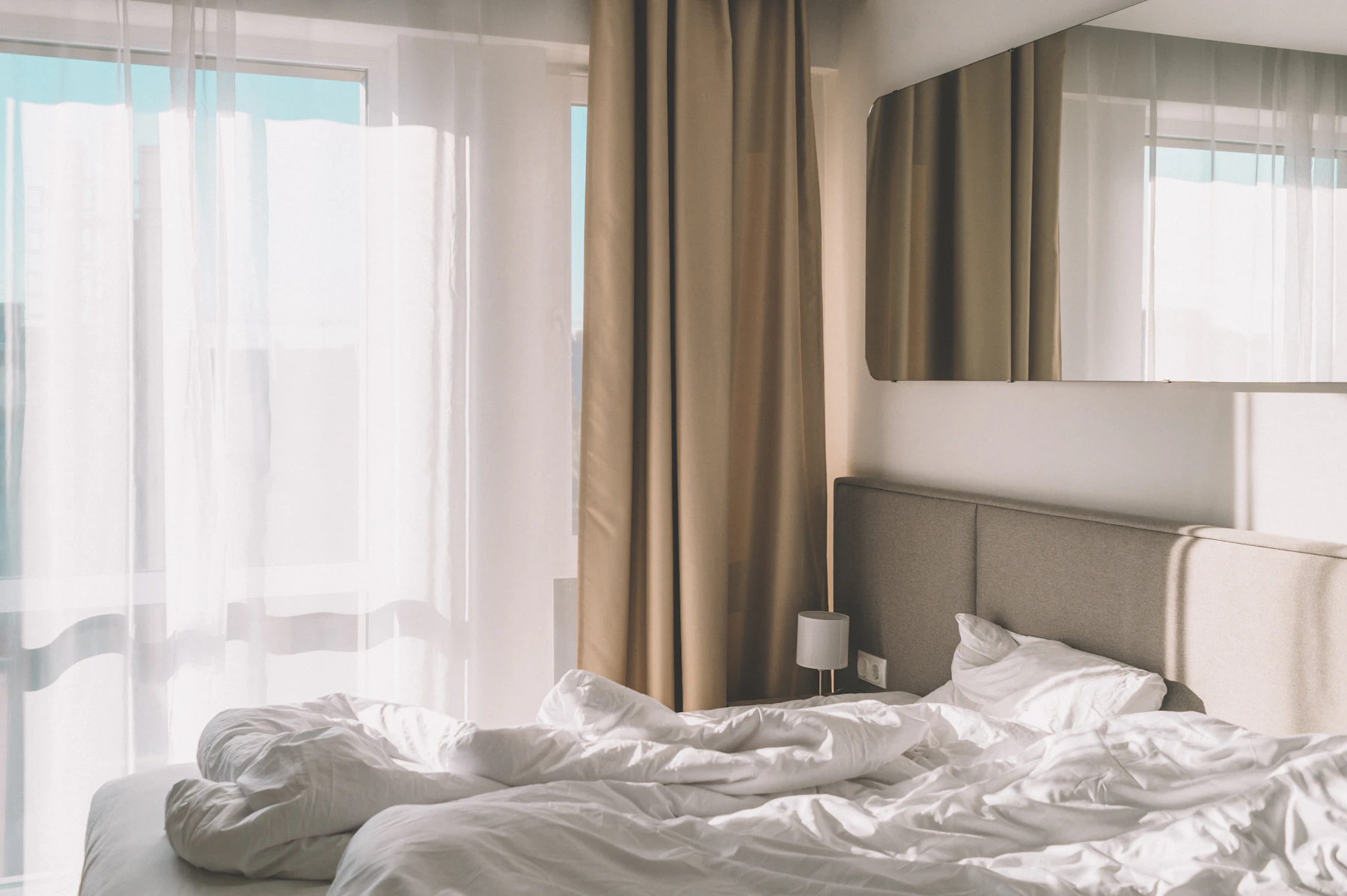 A bright bedroom with white linen, sheer curtains, and a padded headboard