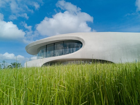A modern, futuristic building with a smooth, curved design is nestled in a vibrant green field under a bright blue sky dotted with fluffy white clouds. Large windows wrap around the upper level, offering panoramic views.