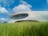 A modern, futuristic building with a smooth, curved design is nestled in a vibrant green field under a bright blue sky dotted with fluffy white clouds. Large windows wrap around the upper level, offering panoramic views.