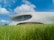 A modern, futuristic building with a smooth, curved design is nestled in a vibrant green field under a bright blue sky dotted with fluffy white clouds. Large windows wrap around the upper level, offering panoramic views.
