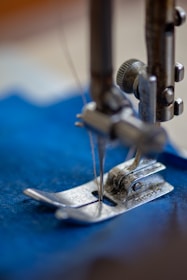 A close-up view of a sewing machine needle and presser foot, partially in focus, working on a piece of blue fabric. The metallic components display a worn and industrial look, suggesting active use.