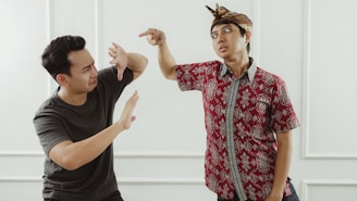 Two individuals are facing each other with expressive gestures. The person on the left is wearing a dark shirt and raising their hands defensively, while the person on the right, dressed in a patterned red shirt and a traditional headpiece, is pointing aggressively.