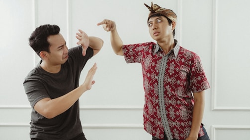 Two individuals are facing each other with expressive gestures. The person on the left is wearing a dark shirt and raising their hands defensively, while the person on the right, dressed in a patterned red shirt and a traditional headpiece, is pointing aggressively.