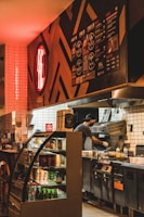 A dimly lit fast-food kitchen with a person preparing food. The interior features a modern design with a red neon sign on the wall. A refrigerated display case with various drinks is visible, and a menu board is mounted above.