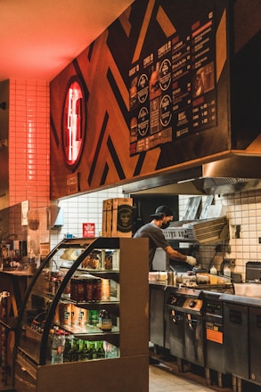 A dimly lit fast-food kitchen with a person preparing food. The interior features a modern design with a red neon sign on the wall. A refrigerated display case with various drinks is visible, and a menu board is mounted above.