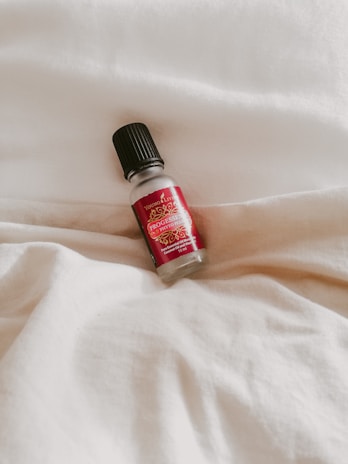 A small bottle with a black cap and red label lies on a white fabric surface. The label contains text and decorative elements.