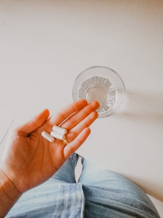 A hand is holding three white capsules above a clear glass of water placed on a light surface. The person is wearing blue denim pants.