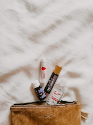 Various small bottles and tubes of personal care products are placed on a soft, textured white surface. These include a grapefruit lip balm, a tranquil roll-on, a valor essential oil, and a Thieves hand purifier, positioned around a brown suede pouch with a zipper labeled 'brownie'.