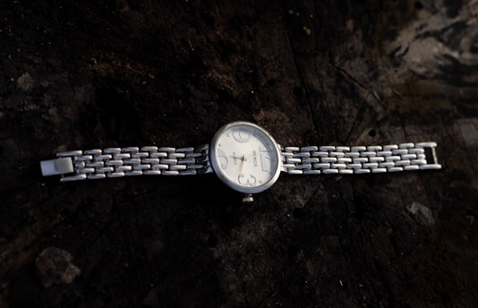 A silver wristwatch with a linked metal bracelet band, resting on a dark, textured wooden surface. The watch face is round with simple hour markers and a minimalistic design.